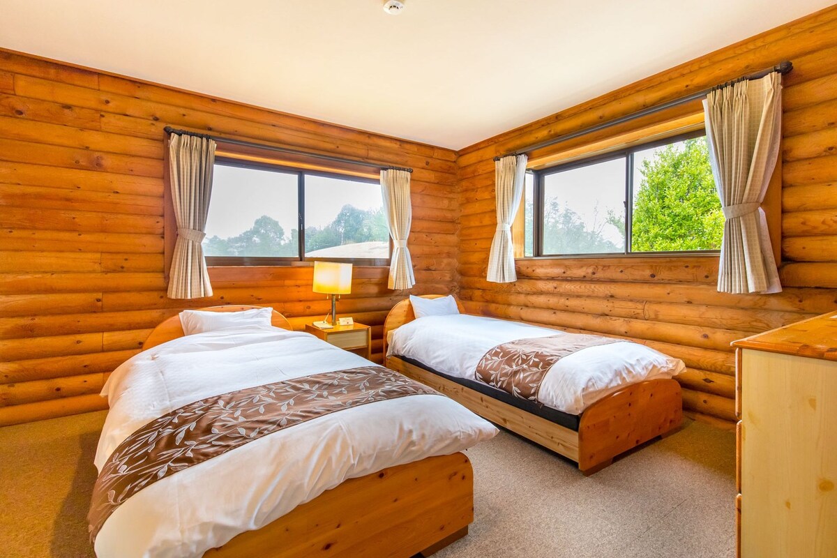 A log cabin bedroom with two beds, a nightstand, a lamp, and a dresser.