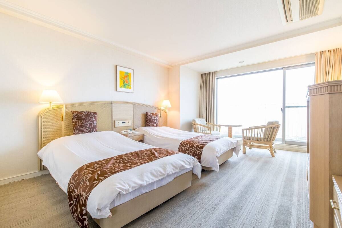 A hotel room with two beds, a small table, a chair, and a large window.