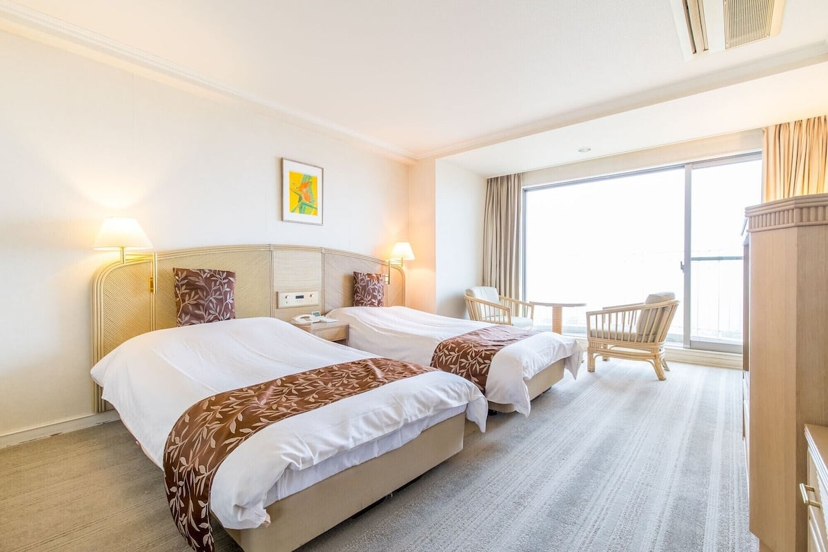 Une chambre d’hôtel avec deux lits, une petite table, une chaise et une grande fenêtre.