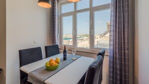 Un coin repas avec une table dressée pour deux personnes, un saladier de fruits, une bouteille de vin et un verre. La pièce est dotée d’une grande fenêtre avec des rideaux, et offre une vue sur la mer et les bâtiments environnants.