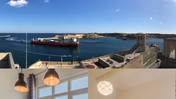 Une vue urbaine sur la côte, avec un grand navire amarré dans le port, un coin repas avec une table et des chaises, et une chambre à coucher avec un lit et des tables de chevet.