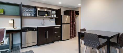 A modern kitchen with a dishwasher, microwave, and refrigerator. There is a dining table with chairs and a lamp.