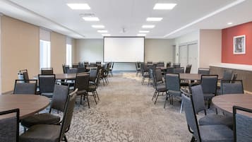Un salón de conferencias con varias mesas y sillas, una pantalla grande y suelo alfombrado.