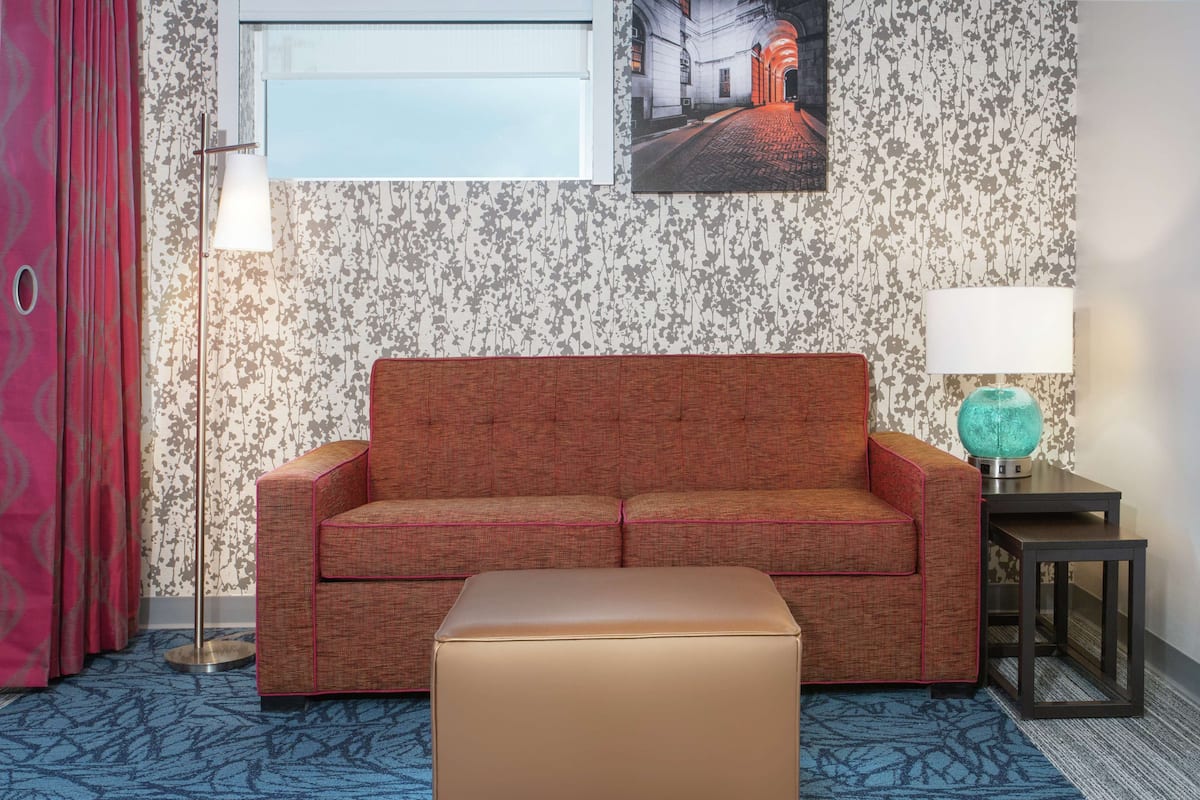 A hotel room with a red sofa, a small ottoman, a floor lamp, a table lamp, and a framed picture on the wall.