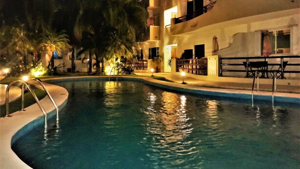 A nighttime view of a swimming pool with a curved edge, surrounded by a building with balconies and outdoor seating. - Condo In Downtown of Playa del Carmen, four blocks to the 5th (Playa del Carmen)
