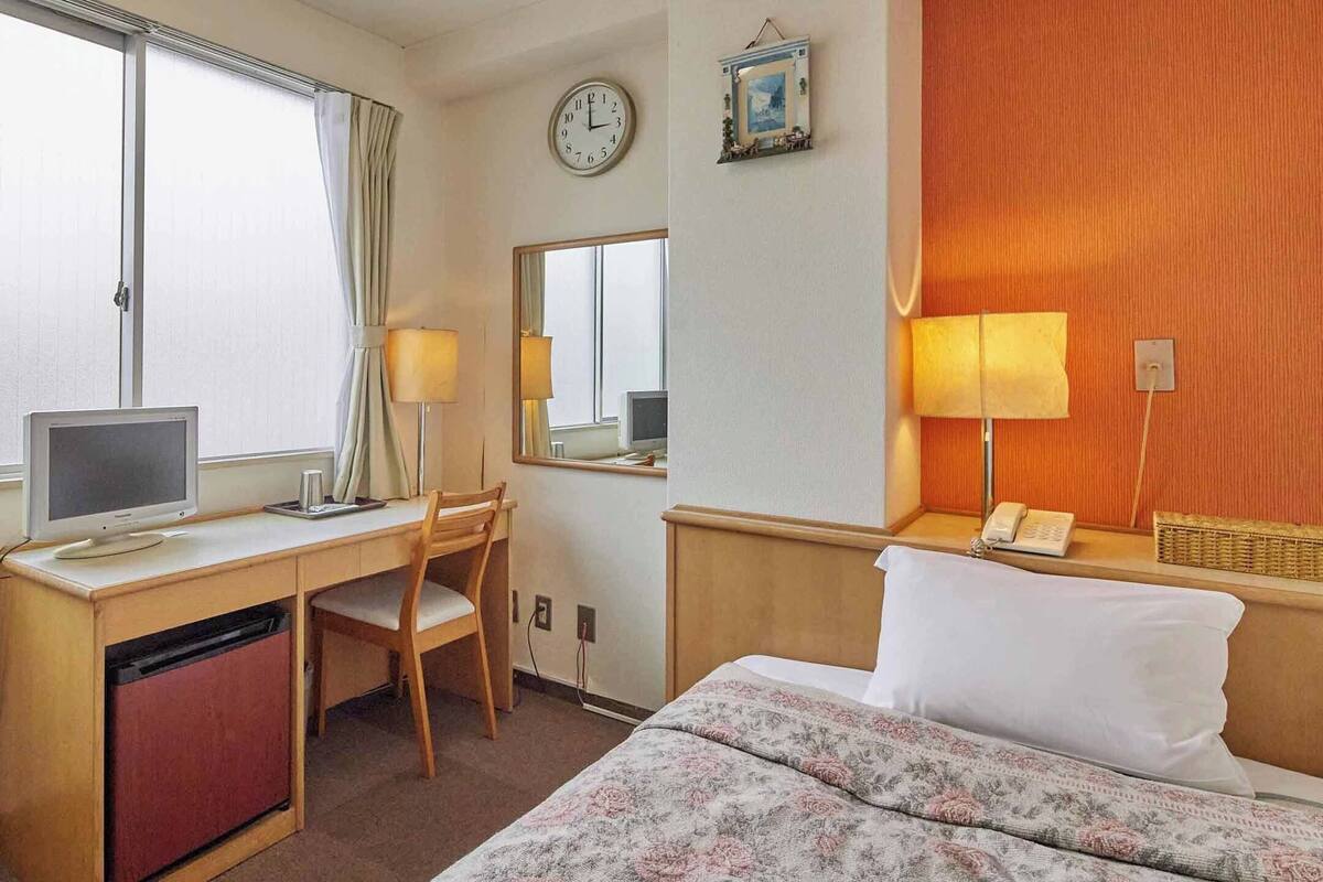 A hotel room with a bed, desk, chair, computer, lamp, clock, and a small bedside table with a phone and a basket.