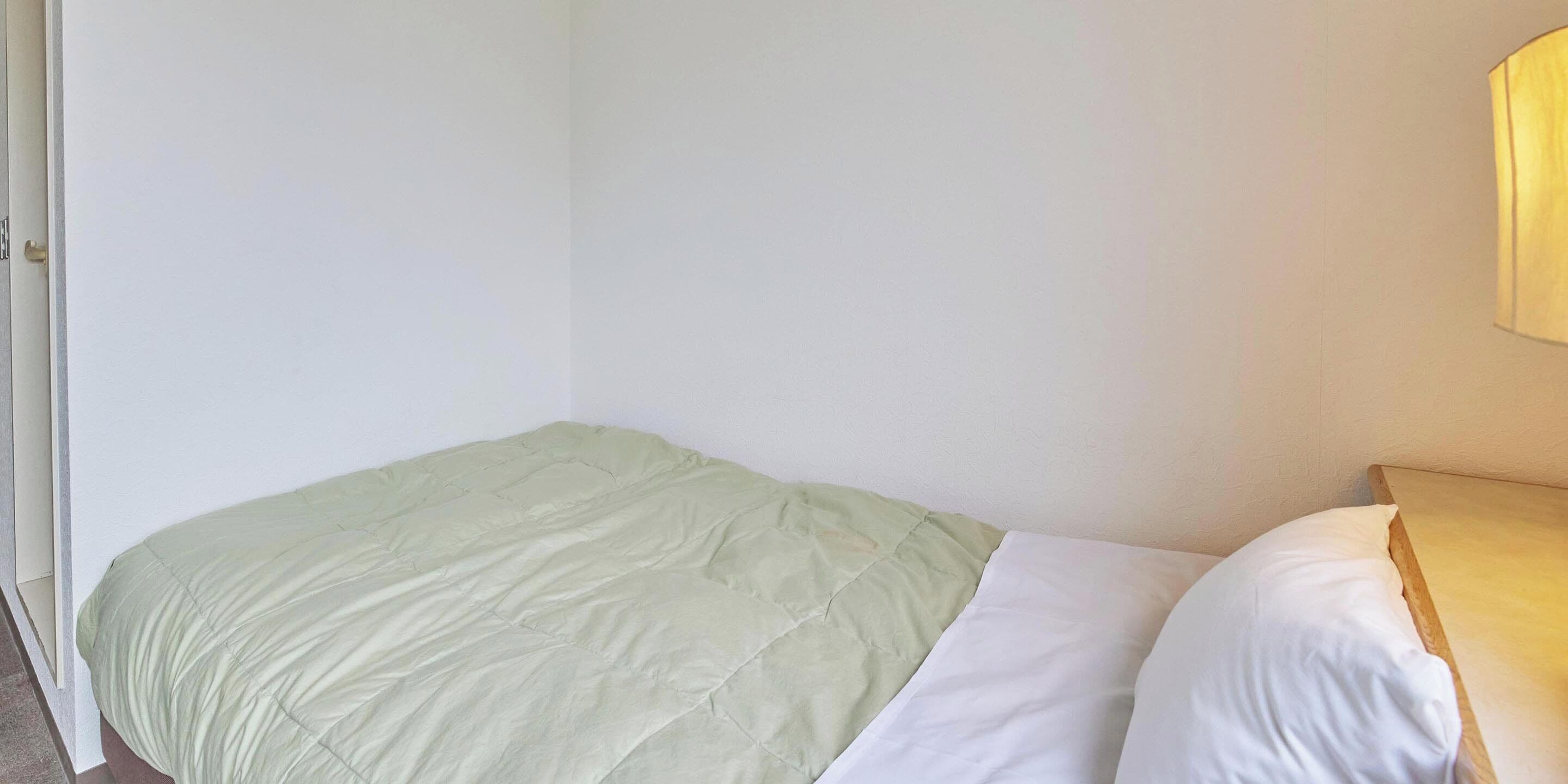 A small, single-bed room with a green bedspread and white pillows, a wooden nightstand, and a wall-mounted lamp.