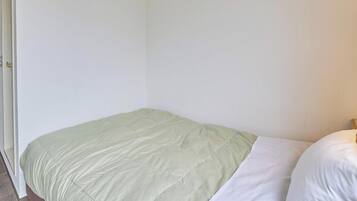 A small, single-bed room with a green bedspread and white pillows, a wooden nightstand, and a wall-mounted lamp.