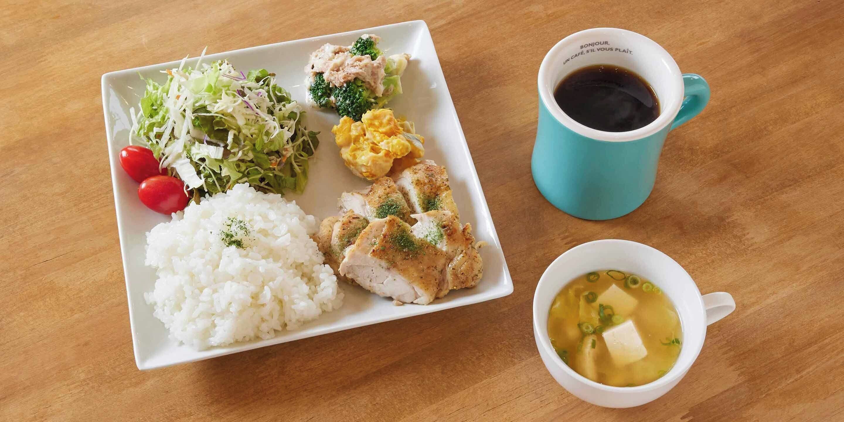 Un repas composé de riz, de poulet, de salade et de soupe, servi dans une assiette blanche, avec une tasse de café bleue à côté.