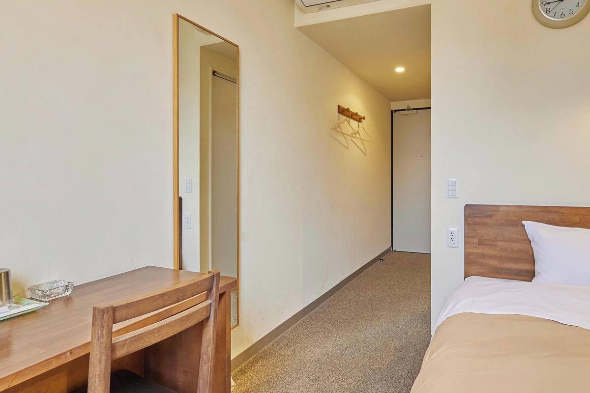 A hotel room with a wooden desk, a bed with a wooden headboard, a wall-mounted clock, and a light fixture.