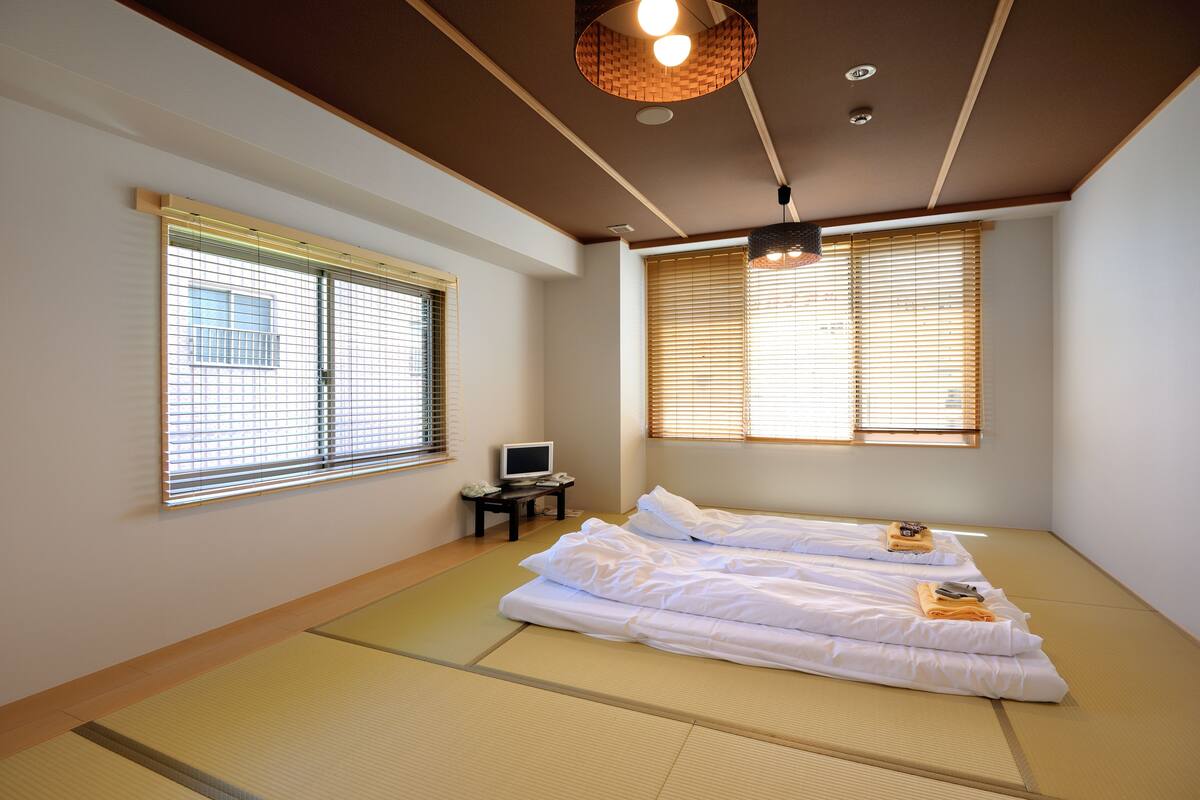 A room with tatami mats, a bed, a television on a small table, and two windows with blinds.