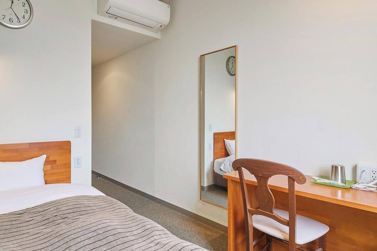 A hotel room with a wooden bed, a desk with a chair, a wall clock, and a mirror.