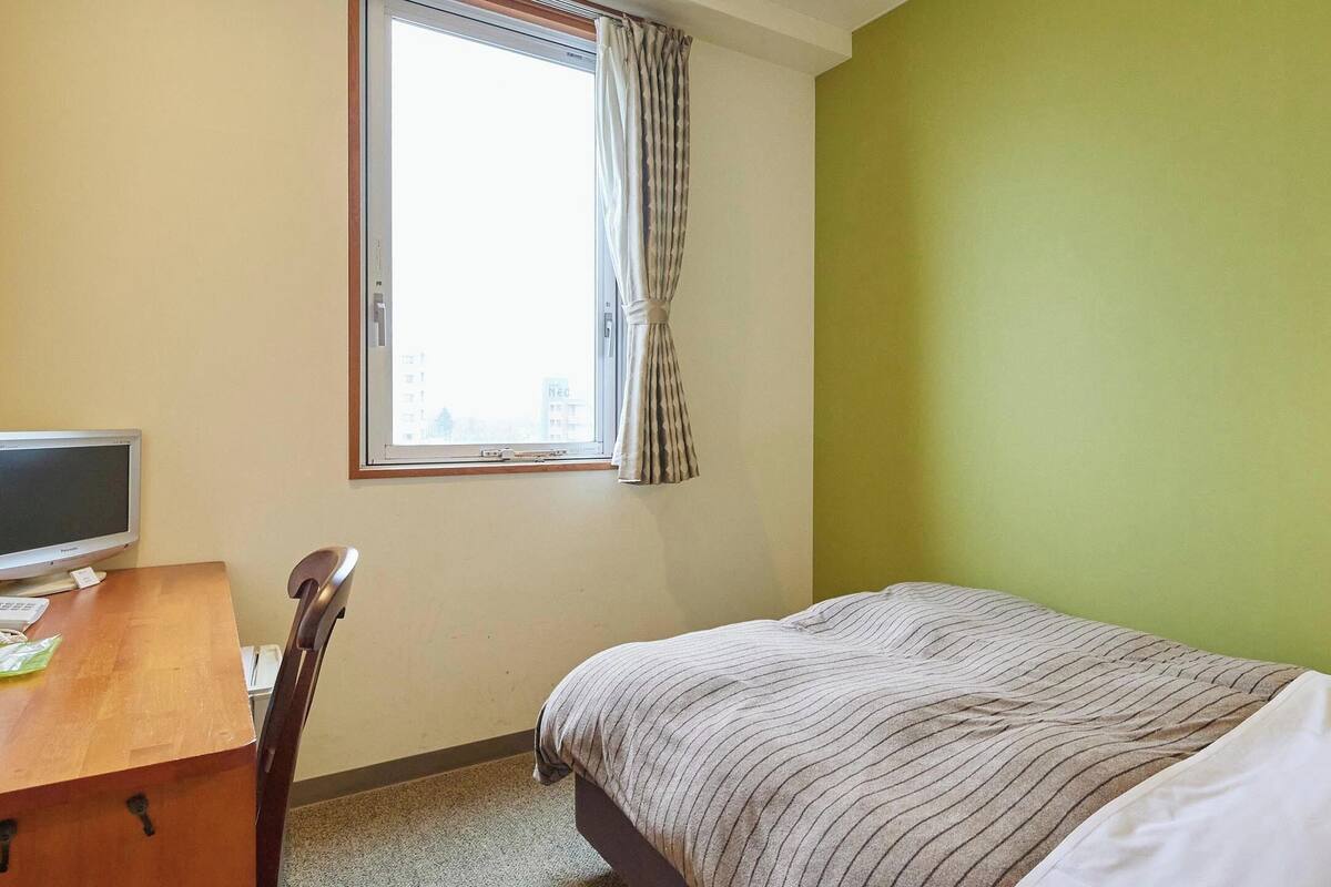 A bedroom with a bed, a desk with a computer, and a window with curtains.
