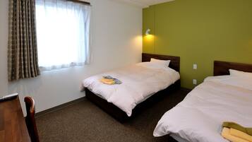 A hotel room with two beds, a wooden desk, a chair, a window with curtains, and a wall-mounted light.