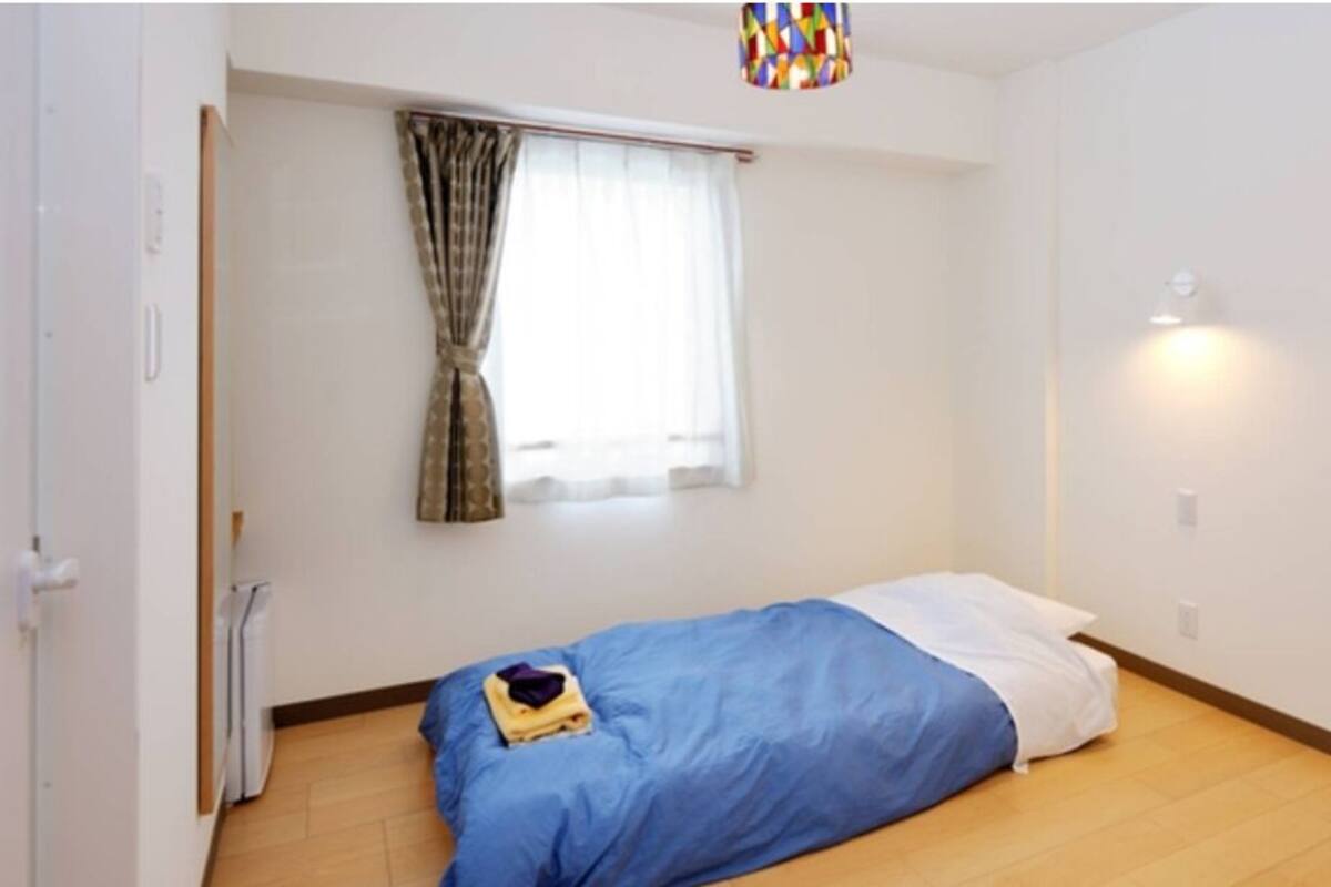 A small, single-bed room with a blue bedspread, a white pillow, and a small table with items on it. There is a window with curtains, a radiator, and a wall-mounted light fixture.