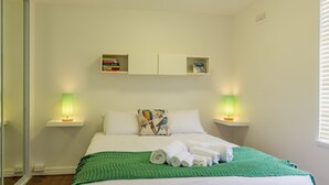 Una cama bien hecha con una manta verde y toallas enrolladas, dos lámparas de noche, una estantería con libros y una almohada estampada en la cabecera.