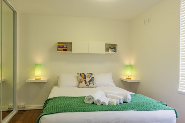 A neatly made bed with a green blanket and rolled towels, two bedside lamps, a bookshelf with books, and a floral pillow on the headboard. - Arwen's Abode by Salamanca (Battery Point)