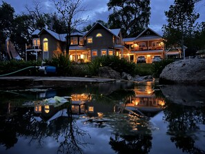 Une grande maison à plusieurs étages, avec un étang devant, entourée d’arbres et d’un jardin bien entretenu.