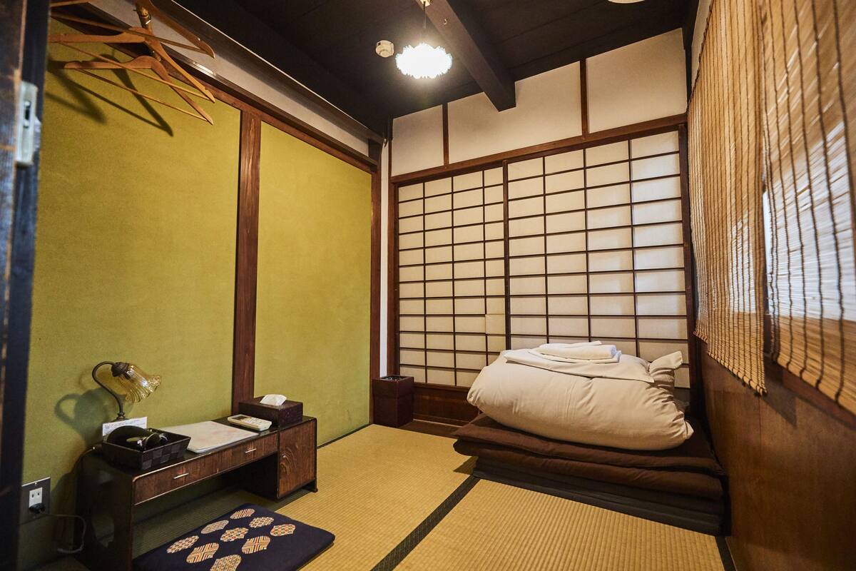 Um quarto tradicional japonês, com piso de tatame, cama de madeira baixa, um painel shoji e uma pequena mesa com abajur e caixa de lenços.
