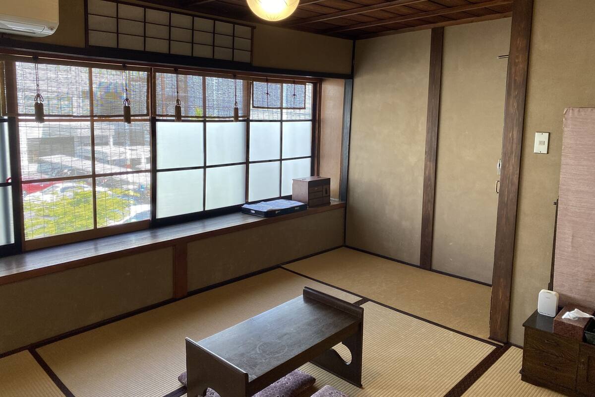Um quarto japonês tradicional com piso de tatame, uma mesa de madeira baixa, uma janela com persianas shoji e um ventilador de teto.