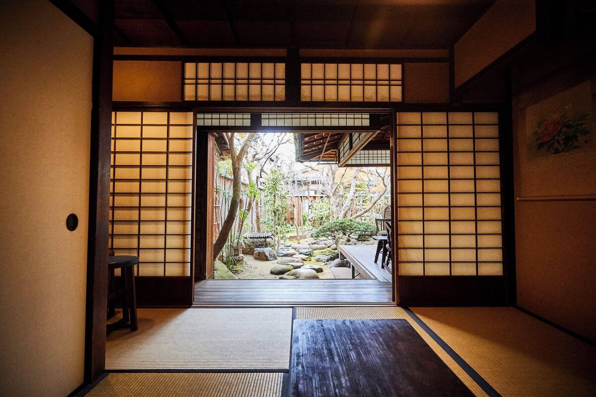 Um quarto japonês tradicional com tatames, uma porta de papel shoji e vista para um jardim com pedras e uma ponte de madeira.