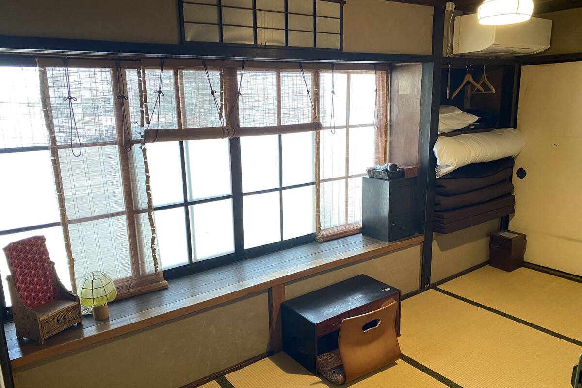Um quarto japonês tradicional com tatames, um futon, um pequeno baú de madeira e uma janela com persianas de bambu.