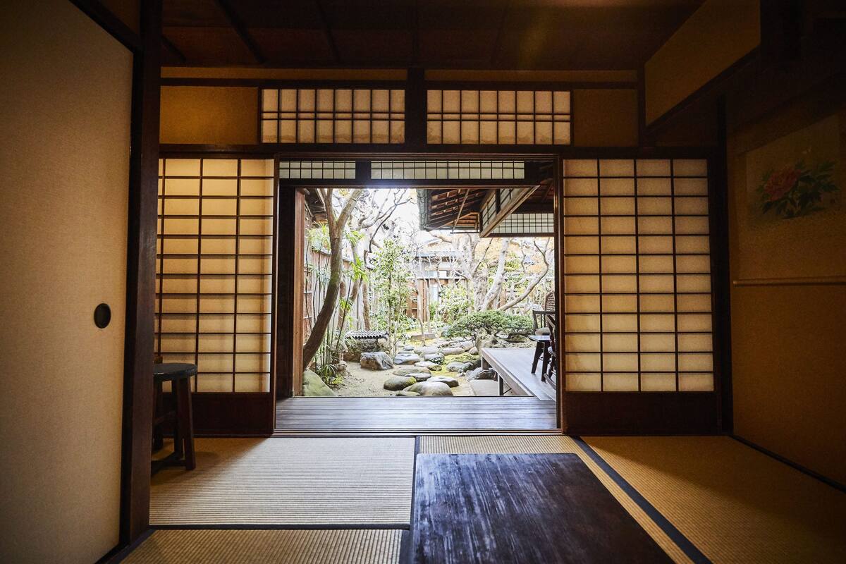 Um quarto japonês tradicional com tatames, uma porta de papel shoji e vista para um jardim com pedras e uma ponte de madeira.