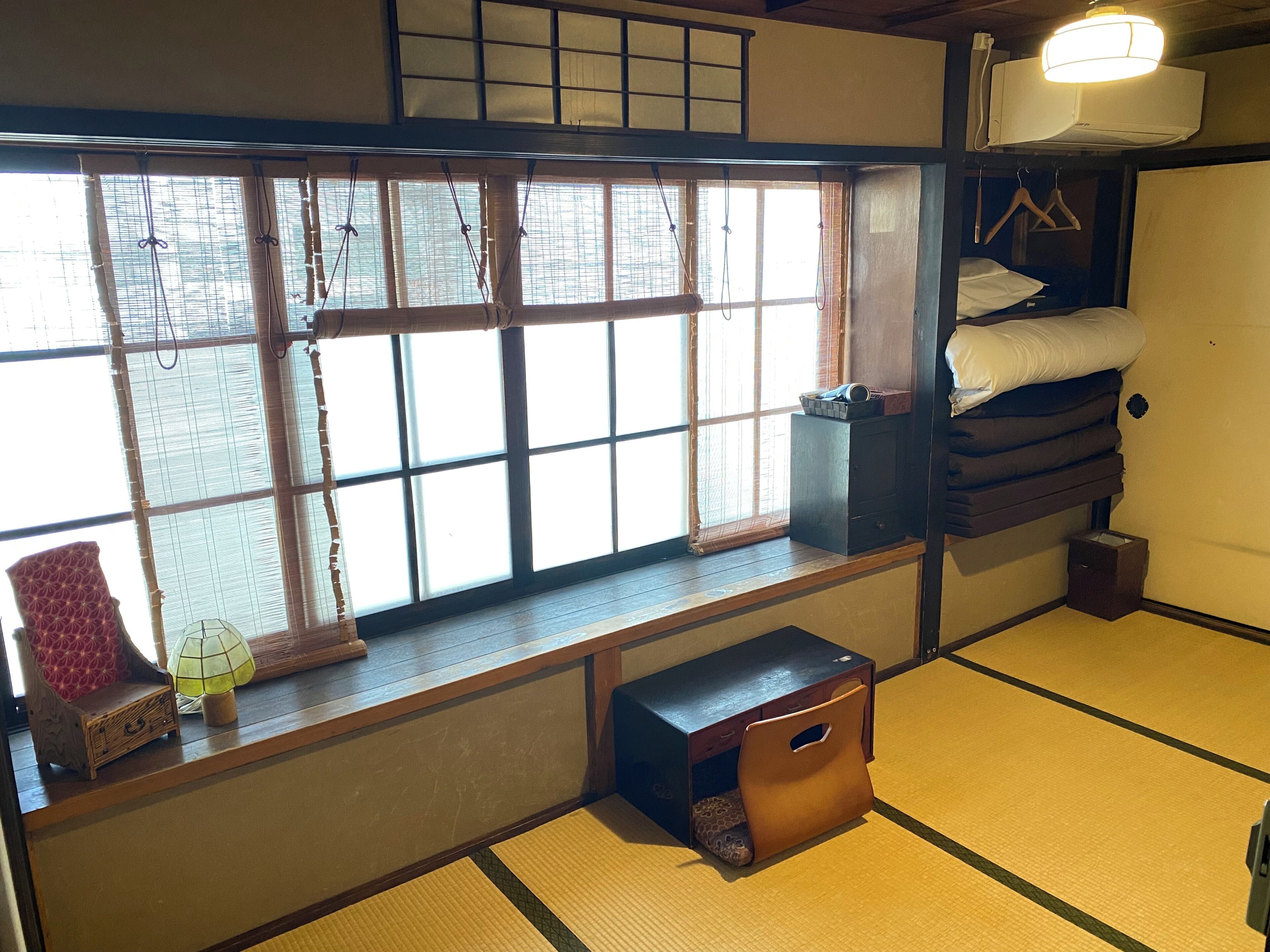 A traditional Japanese room with tatami flooring, a futon bed, a small wooden chest, and a window with bamboo blinds.