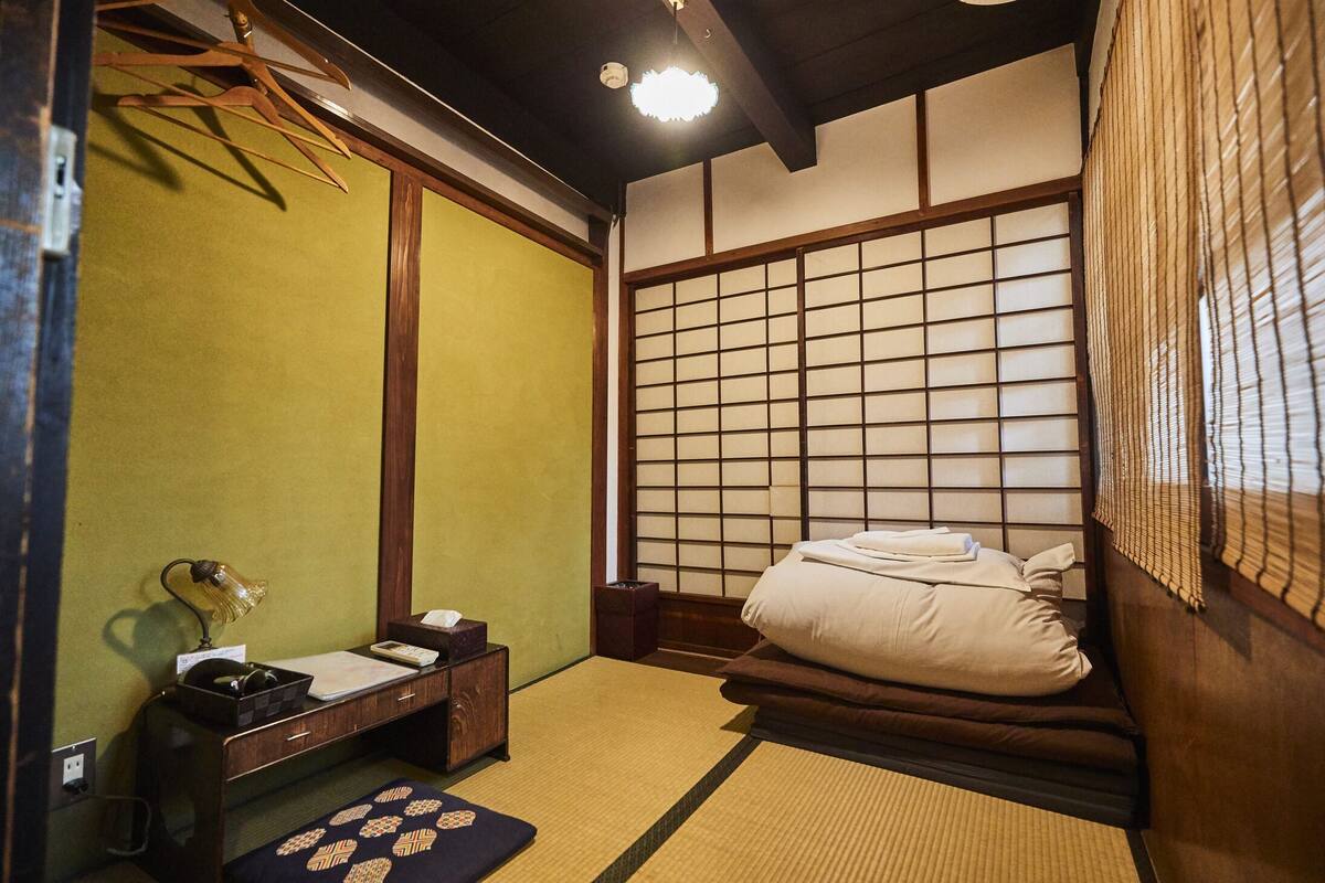 Une pièce de style japonais traditionnel, avec un sol recouvert de tatamis, un futon servant de lit, un paravent en papier de riz (shoji) et un petit bureau avec une lampe.