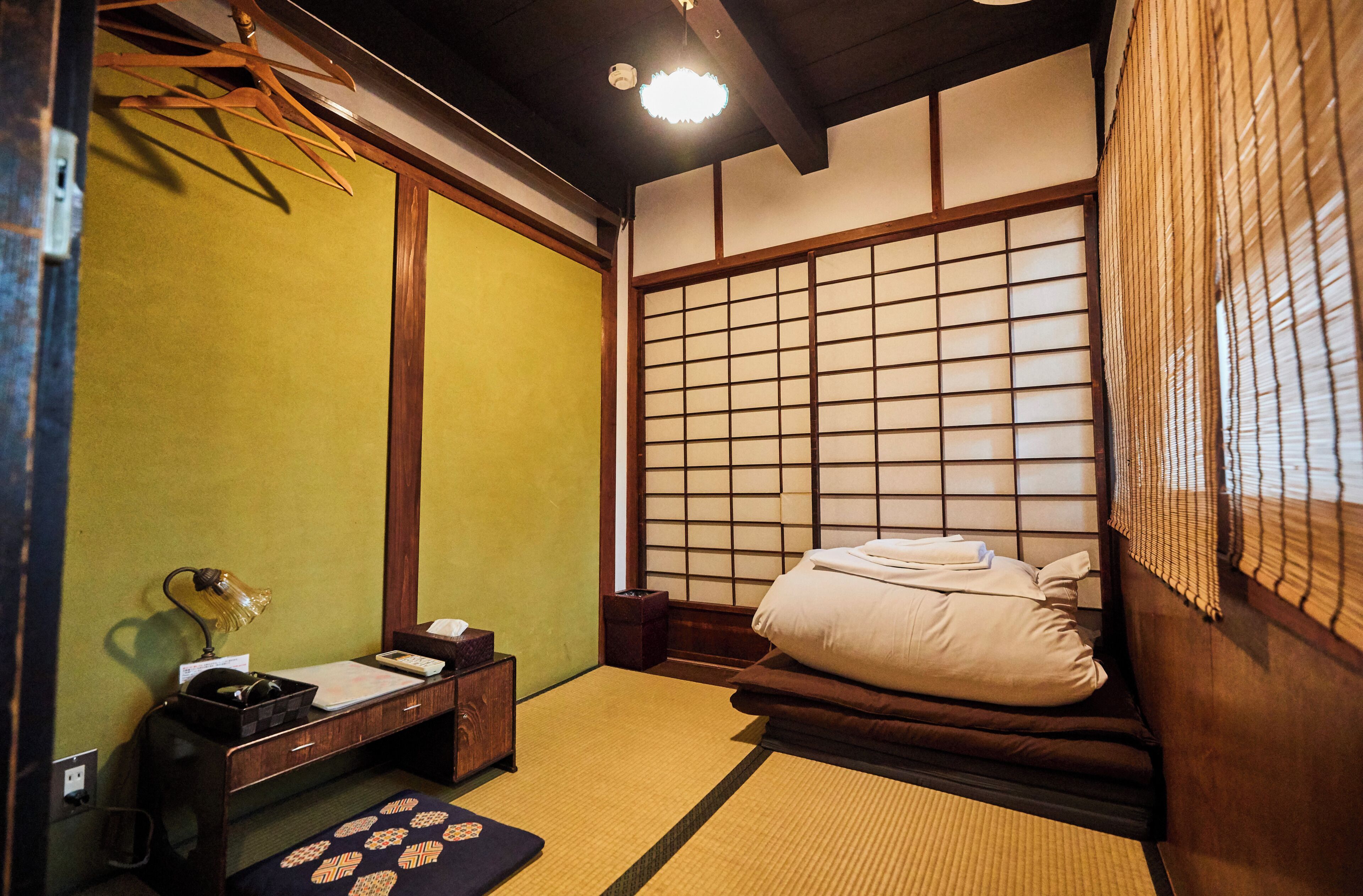 A traditional Japanese-style room with tatami flooring, a low wooden bed, a shoji screen, and a small desk with a lamp and a tissue box.