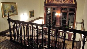Una gran escalera con barandales ornamentados, un amplio arco de entrada con un detallado cuadro, y obras de arte enmarcadas en las paredes.