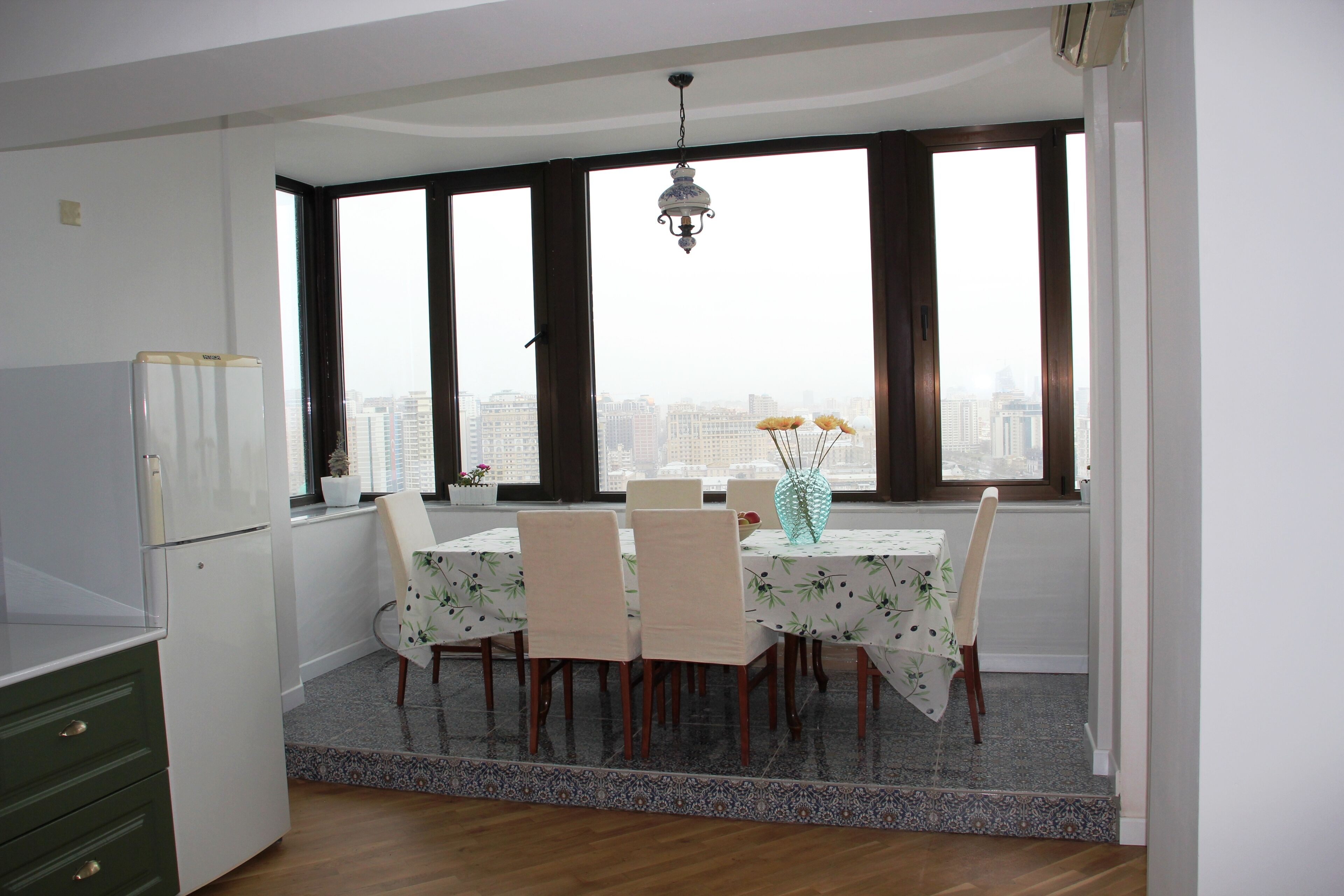 A dining area with a table set for four, a refrigerator, and a view of the city through large windows.