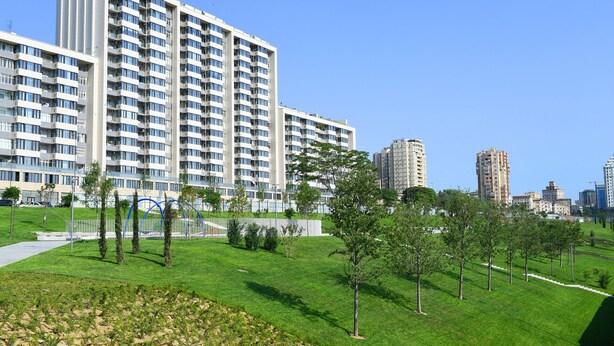 A modern apartment building with multiple floors and balconies, surrounded by well-maintained green spaces and trees.