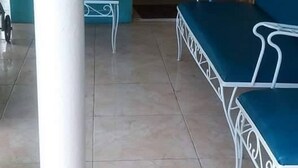Uma área de estar coberta ao ar livre com duas cadeiras azuis e um banco, um pilar branco e um tapete de entrada.