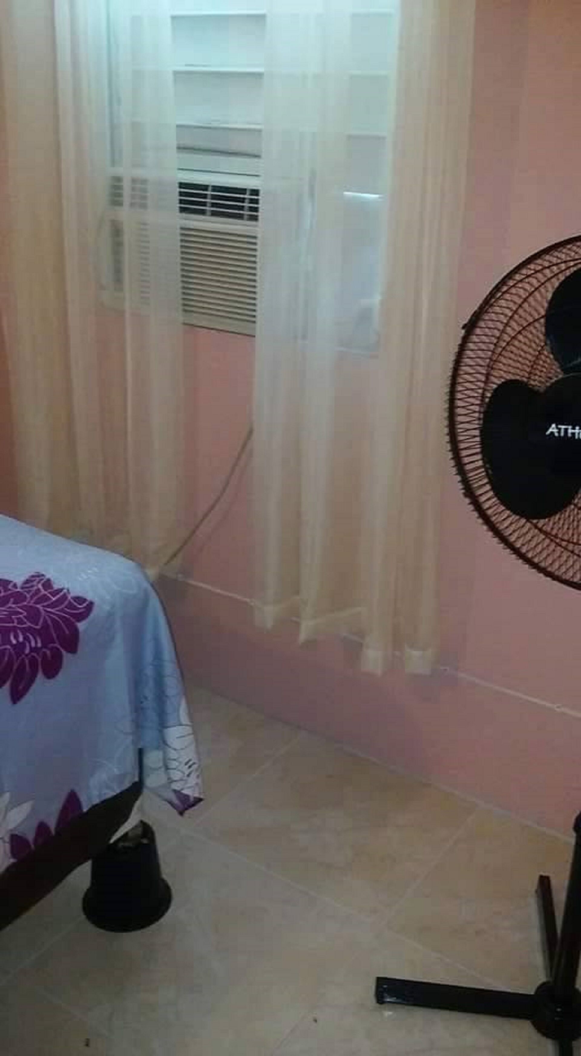 A bedroom with a bed, a window with sheer curtains, a wall-mounted fan, and a radiator.