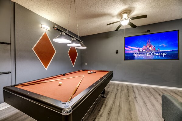 A pool table in a room with a ceiling fan, a wall-mounted TV displaying a Disney movie, and a geometric wall decoration.