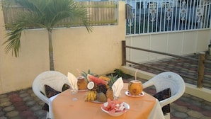 Un espace de restauration en plein air avec une table dressée pour deux personnes, agrémentée d’un plateau de fruits, d’une noix de coco et d’un verre rempli d’une boisson.