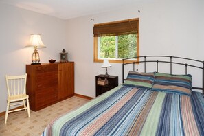A bedroom with a bed, a wooden dresser, a chair, a nightstand, a lamp, and a window with a view of greenery.