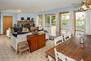 A spacious living room with a fireplace, a dining area with a wooden table and chairs, and large windows offering a view of the outdoors.