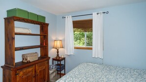 Ein Schlafzimmer mit einem hölzernen Schminktisch, einem Nachttisch mit Lampe, einem Fenster mit Vorhängen und einer blumigen Bettdecke.
