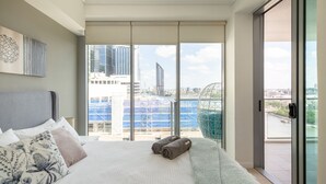 Une chambre d’hôtel moderne dotée d’un grand lit, offrant une vue sur la ville grâce à de grandes fenêtres du sol au plafond, et d’un balcon avec une rambarde en verre.