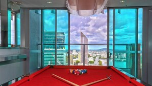 Une table de billard recouverte d’un drap de feutre rouge, une bille blanche et plusieurs billes de couleurs différentes disposées en forme de triangle. La pièce est dotée de grandes fenêtres qui offrent une vue sur un paysage urbain avec des bâtiments modernes et un drapeau.