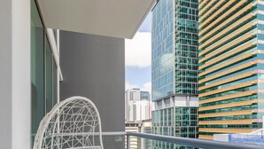Un balcon avec une chaise en osier blanc et un coussin bleu, d’où l’on peut admirer un paysage urbain avec des bâtiments modernes.