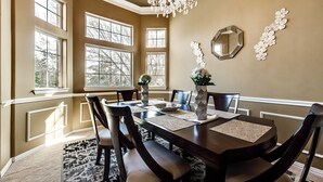 Ein Esszimmer mit einem dunklen Holztisch, sechs Stühlen und einer Chandelier. Große Fenster sind an der linken Seite, und ein Spiegel ist an der Wand angebracht.