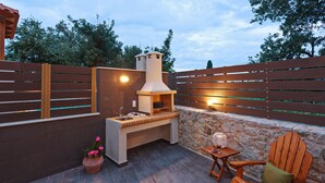En udendørs terrasse med en stenvæg, grill i træ, en træstol og et lille bord med en lampe.