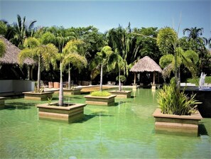 A pool area with clear water, surrounded by palm trees and thatched-roof structures.