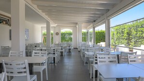 Uma ampla área de refeições interna com mesas e cadeiras brancas, grandes janelas com vista para o verde e teto com vigas aparentes.