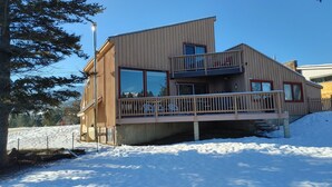 A modern wooden cabin with a balcony, large windows, and a snow-covered ground.