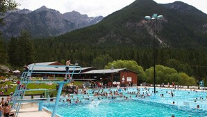 A swimming pool with a diving platform, surrounded by mountains and a small building.