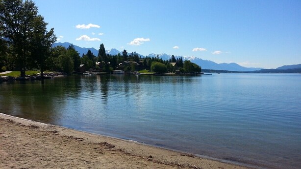 A lake with a sandy beach, trees, and houses in the background.